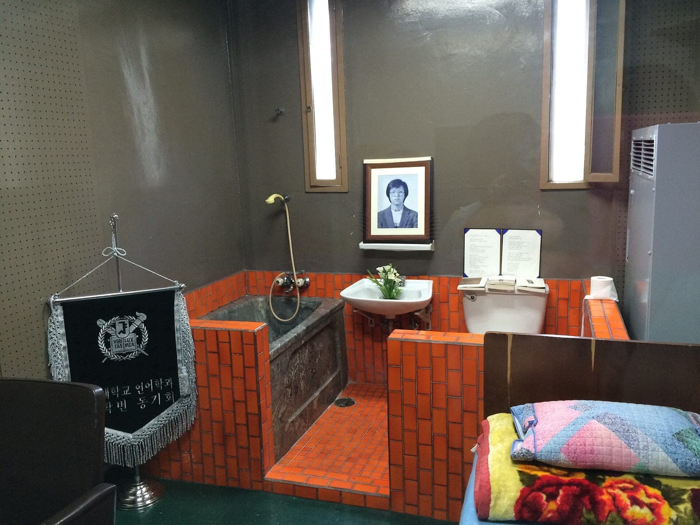 A small tiled room with a bathtub, a washbasin, a narrow bed and a framed portrait of Park Jong-chul on the wall.